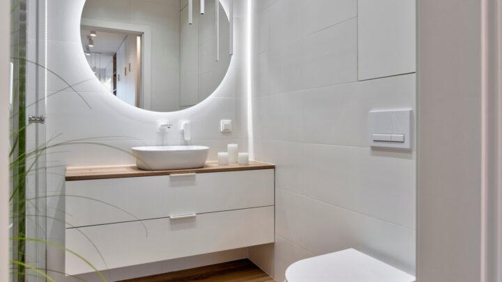 Innenraum eines stilvollen Badezimmers mit Keramikschale auf dem Holzschrank, Holzfliesen auf dem Boden, runder Spiegel mit LED-Leuchten.