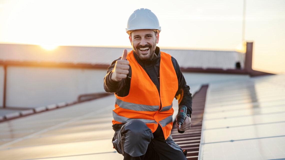 Ein glücklicher Arbeiter mit Bohrmaschine in den Händen gibt Daumen hoch für die Verwendung von Sonnenkollektoren.