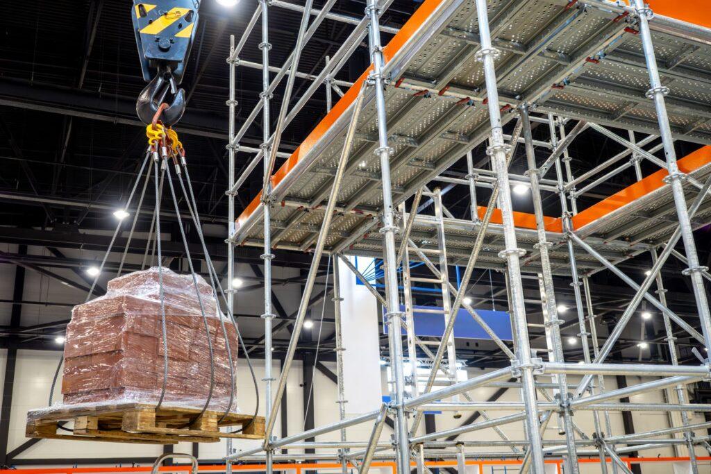 Baustellenszene mit Kranhaken, der eine Palette mit Baumaterialien an einem Gerüst hebt